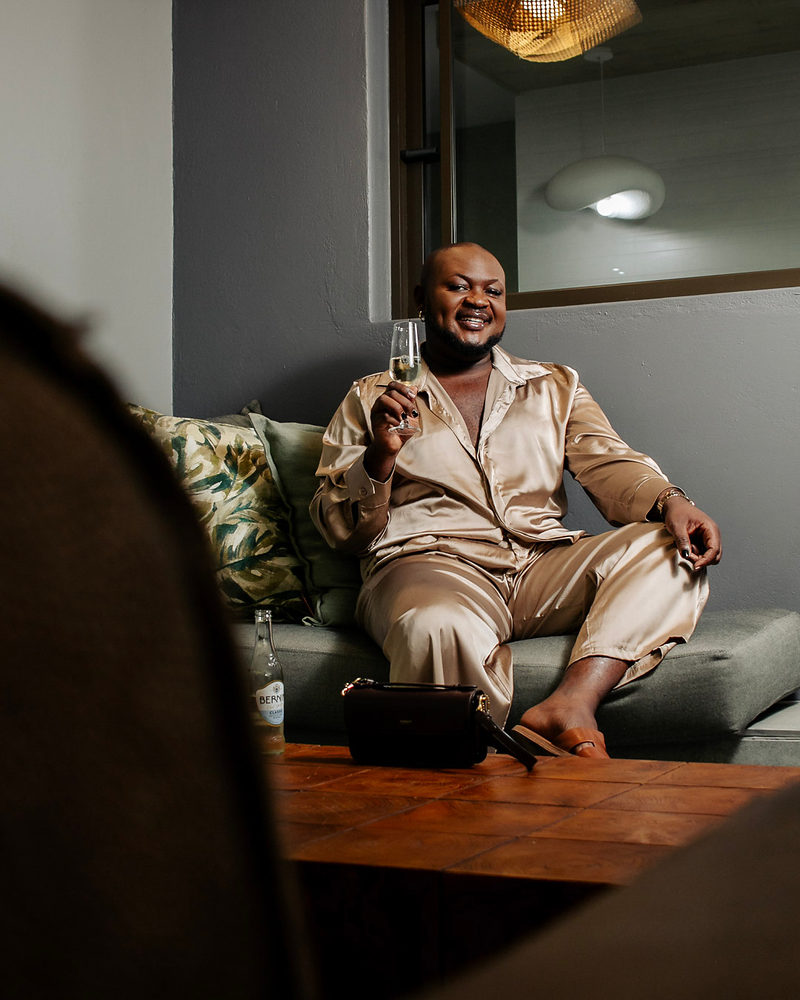 A smiling man in a champagne satin set holding a champagne flute, seated on a green sofa